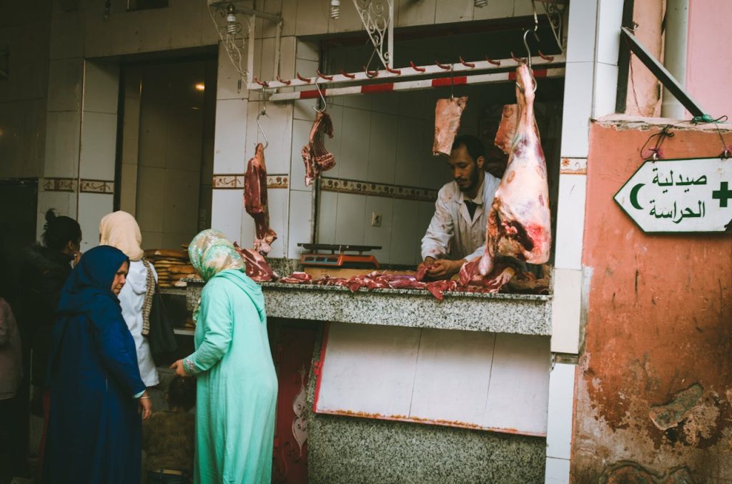A lively outdoor market scene showing a butcher serving customers at a meat stall.